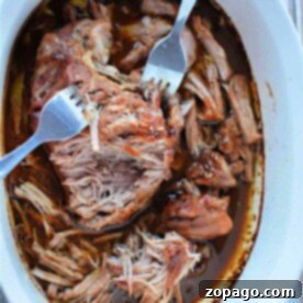 Succulent shredded pork in a white oval dish awaits, accompanied by two forks for easy serving. Master the art of cooking pulled pork and savor its tender goodness.