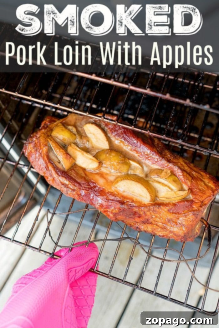 Amazing apple stuffed smoked pork loin recipe that takes just 15 minutes to prep! With our dry rub and fruit, you'll have tender pork you'll love. #smoked #pork #easy #dryrub #apple #best #electricsmoker #smokerrecipes #smokedpork A beautifully smoked pork loin, artfully topped with caramelized apple slices, resting on a grill rack. A pink oven mitt gently supports the rack, highlighting the golden-brown exterior and succulent texture of this exquisite apple stuffed smoked pork recipe.