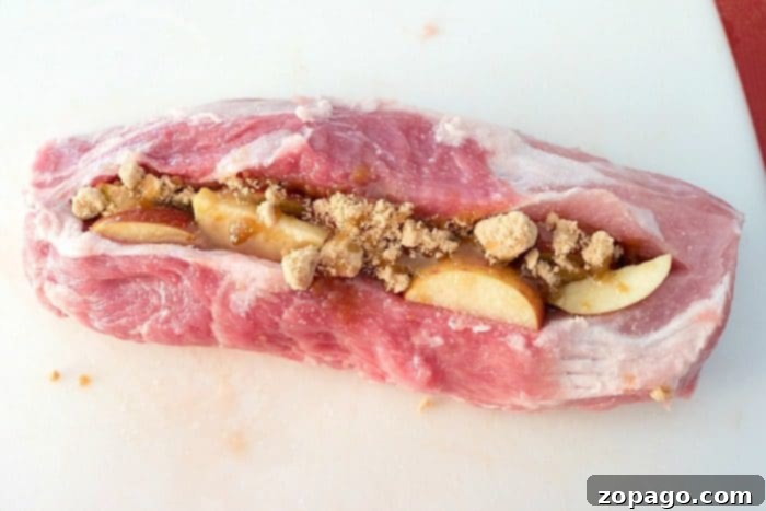 Discover the most amazing apple stuffed smoked pork loin recipe, with just 15 minutes of prep! Our dry rub and fruit guarantee tender pork you'll adore. #smoked #pork #easy #dryrub #apple #best #electric A pristine raw pork loin, meticulously prepared for smoking, is showcased on a white cutting board. It's carefully slit and generously stuffed with fresh apple slices and crumbled brown sugar, illustrating a crucial step for this delightful smoked pork recipe.