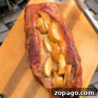 A perfectly smoked pork loin, stuffed with tender apple slices, presented on a wooden cutting board, ready to be carved. Tongs rest beside it, showcasing this exquisite smoked pork recipe.