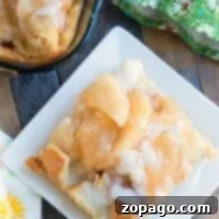 A plate of apple pie bread pudding with glaze rests beside a green floral napkin and a baking dish.