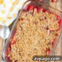 A glass baking dish filled with a cranberry apple cobbler, featuring sliced apples and cranberries topped with a golden oat crumb layer. It is placed on a wooden surface, surrounded by a spoon, a yellow-patterned cloth, an apple, and an orange slice.