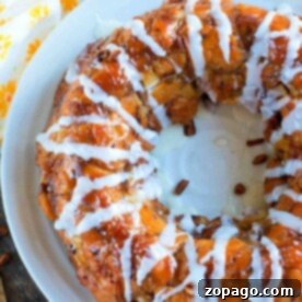 A round pastry drizzled with white icing sits on a white plate, evoking the cozy delight of apple monkey bread made from cinnamon rolls. Juicy apples peek from the background, completing the scene.