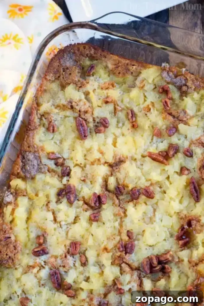 A close-up of a Gingerbread Dump Cake in a glass dish. The topping consists of chopped pecans and pieces of pineapple, creating a golden-brown crust. The edges are slightly crisp, and a patterned yellow and white towel is partially visible beside the dish.