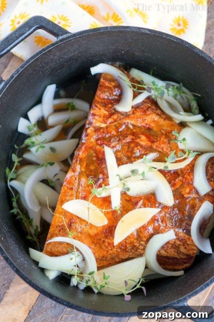 This Dutch oven ham with a honey glaze is a must-try for your holiday dinner, making it one of the best Dutch oven recipes. A seasoned ham with sliced onions and herbs in a Dutch oven, ready for baking.