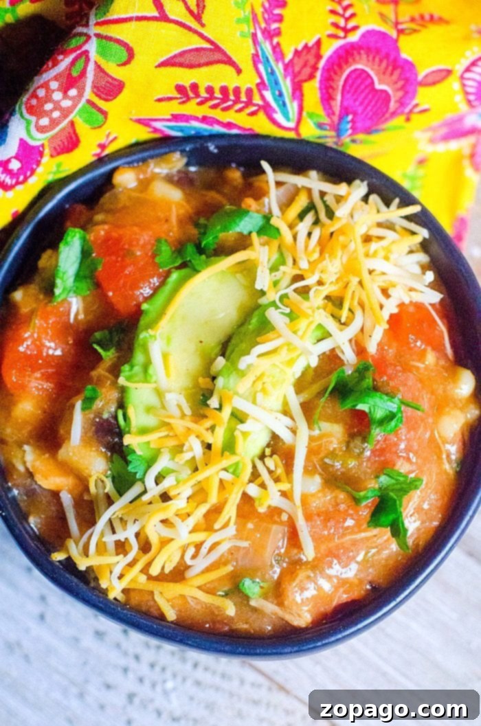 This Instant Pot turkey soup is a great leftover turkey recipe that's thick and flavorful like an enchilada soup packed with protein! You've gotta' try it. #instantpotrecipes #instantpot #leftoverturkey #turkey #soup #enchilada #thetypicalmom A bowl of Instant Pot turkey soup topped with sliced avocado, shredded cheese, and chopped cilantro. The bowl rests on a wooden surface next to a colorful, patterned fabric. The soup features chunks of vegetables in a rich tomato-based broth.