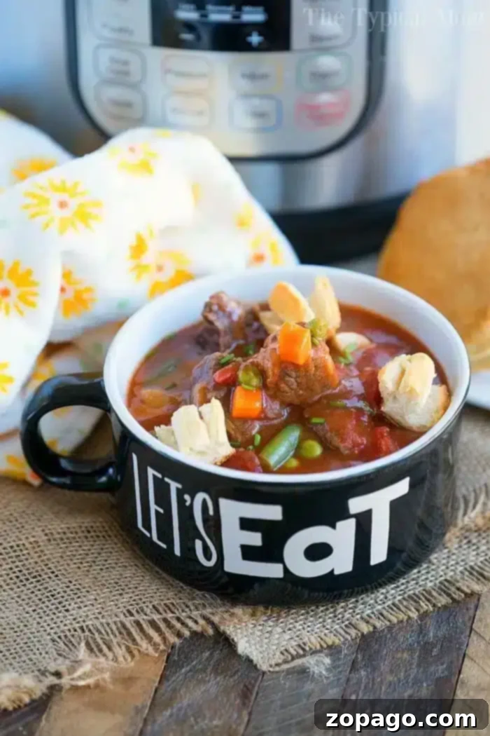 Instant Pot Beef Pot Pie Soup - Your Family's New Favorite Hearty Dinner A black mug emblazoned with 'Let's Eat' brims with pressure cooker beef pot pie soup, garnished with tiny pie crust shapes. It sits on a wooden surface alongside a white floral cloth, while a pressure cooker lingers in the background.