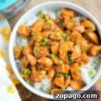 Effortless Instant Pot Sesame Chicken 4 pressure cooker sesame chicken, perfectly glazed and sprinkled with green onions, rests atop a bed of rice on a wooden surface. A floral napkin adds an elegant touch to the setting.