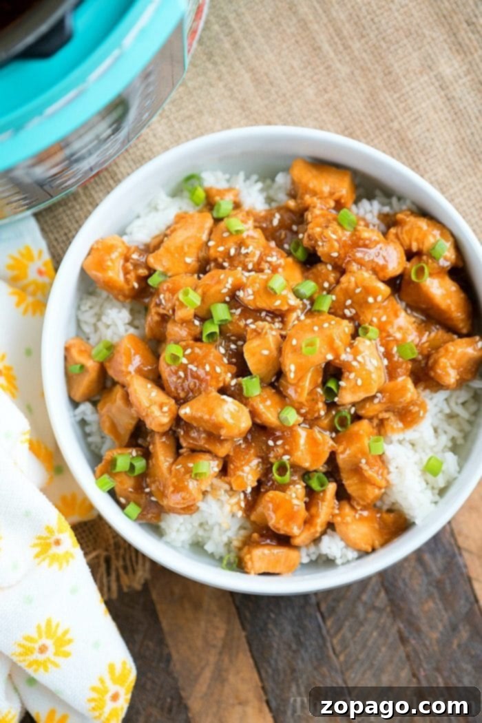 Pressure cooker sesame chicken, perfectly glazed and sprinkled with green onions, rests atop a bed of rice on a wooden surface. A floral napkin adds an elegant touch to the setting.