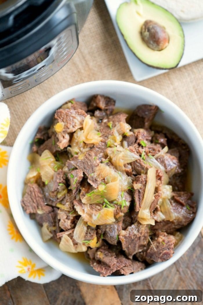 French onion pressure cooker roast is tender with tons of flavor. Perfect Instant Pot shredded beef tacos for dinner even your kids will rave about. A bowl of shredded beef with onions, reminiscent of French onion flavors, is served beside an avocado half on a wooden surface.