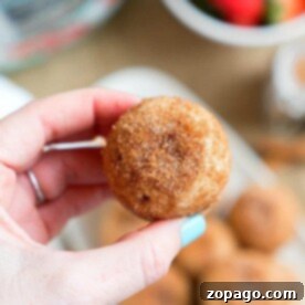 Crispy Instant Pot Churro Bites 3 A hand holds a round, cinnamon-sugar-coated treat, reminiscent of pressure cooker churro bites. In the blurred background, more of these tasty delights sit alongside a bowl brimming with strawberries.