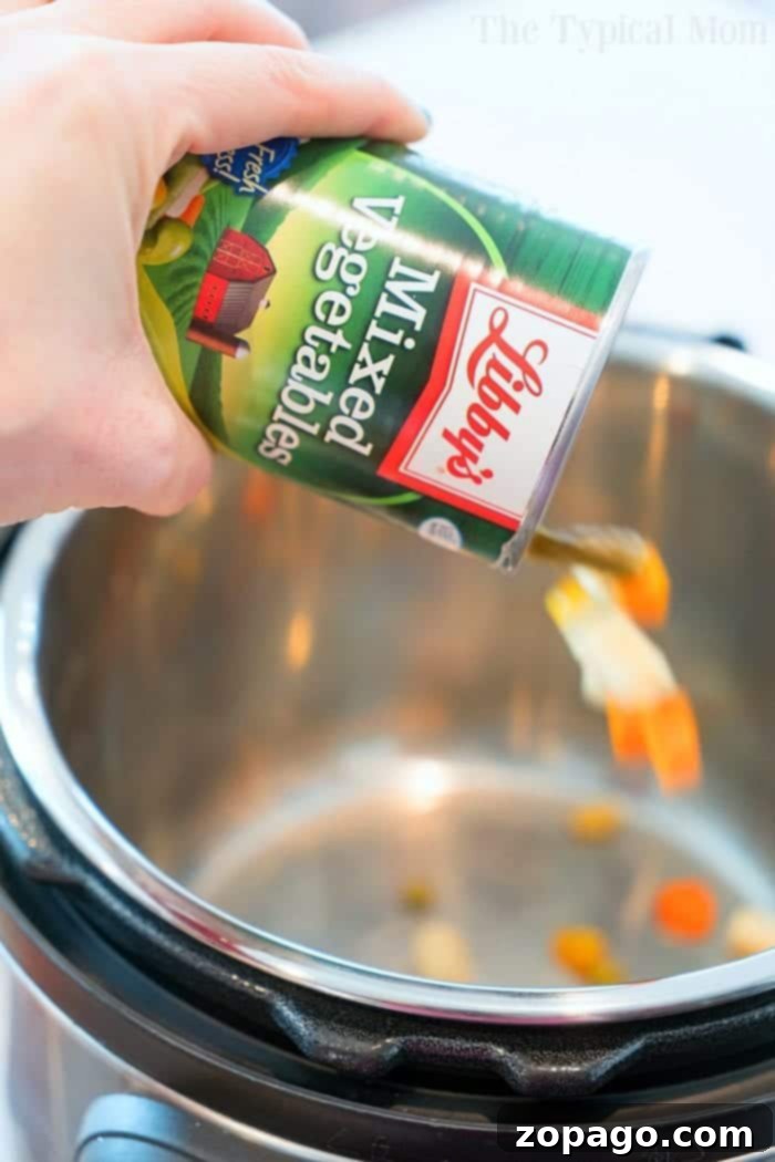Pressure cooker chicken pot pie soup is a spoonful of comfort food made in under 15 minutes! You've got to try this creamy chicken soup with veggies today. #instantpotrecipes #pressurecooker #chickenpotpie #soup #chickensoup #ninjafoodirecipes #crockpotexpressrecipes #thetypicalmom A hand pouring canned mixed vegetables into a pressure cooker for a cozy chicken pot pie soup, highlighting the ease of preparation.