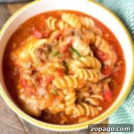 A bowl of rotini pasta, dubbed Ninja Foodi taco pasta, is topped with a chunky tomato and ground meat sauce, garnished with melted cheese and herbs. The bowl rests on a wooden surface beside a green cloth, with part of a kitchen appliance visible in the background.