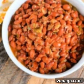A white bowl filled with seasoned pinto beans, fresh from the pressure cooker, sits on a wooden surface. The reddish-brown beans are tantalizingly presented alongside a sliver of floral-patterned cloth peeking in from the top left corner.
