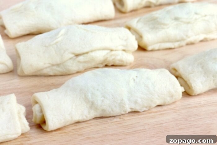 Several unbaked crescent roll dough squares, filled and rolled, arranged on a wooden surface, ready to be fried into peanut butter and jelly donuts.