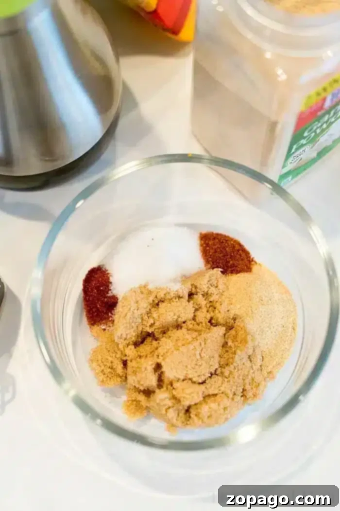 Best air fryer chicken thighs ever! Use this dry rub for chicken to make sweet and crispy chicken in an air fryer that's tender and juicy inside. Yum! #airfryerrecipes #airfryer #ninjafoodirecipes #ninjafoodi #chickenthighs #chicken #crispy A glass bowl containing various seasonings, including brown sugar, garlic powder, paprika, and salt. A metal object and a container of spices are partially visible in the background on the white surface—perfect for flavoring bone-in air fryer chicken thighs.