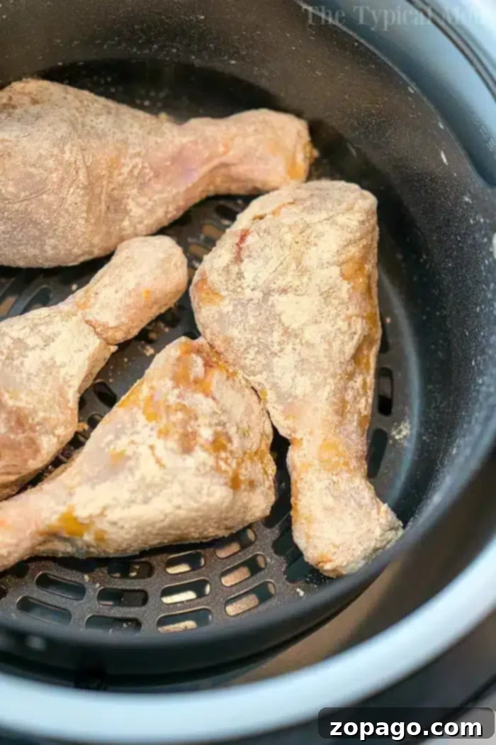 Air Fryer Fried Chicken - Breaded Drumsticks in Basket A close-up view of four raw, breaded chicken drumsticks carefully arranged in the black mesh basket of an air fryer, poised for cooking. The chicken is evenly coated with a light flour mixture, showcasing the simple preparation for an easy air fryer fried chicken recipe. The texture of the drumsticks promises a future crispy exterior.