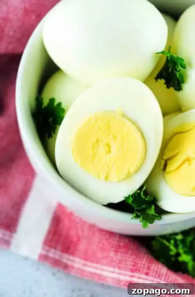 Air Fryer Hard Boiled Eggs 2 A bowl contains several air fryer hard boiled eggs, with one egg cut in half to reveal the vibrant yellow yolk. The bowl is garnished with fresh green parsley and placed on a charming red and white checkered cloth, inviting a delightful culinary experience.
