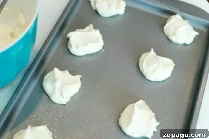 Effortless Angel Food Two-Ingredient Cookies 4 Delicate dollops of white, airy cookie batter resting on a baking sheet, ready for the oven, beside a blue mixing bowl.