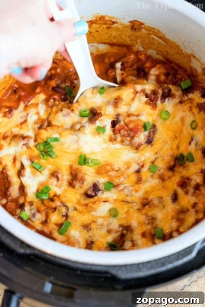 Quick Pressure Cooker Cheesy Chili Mac 4 A hand holding a white spoon is stirring a cheesy, baked pressure cooker chili mac topped with green onions in a pot. The dish appears to have a tomato-based sauce and melted cheese.