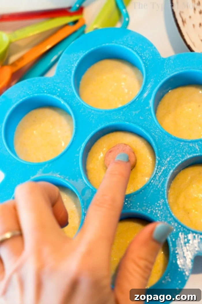 Pressure cooker corn dog bites are easy to make in your Instant Pot, Crock Pot Express or Ninja Foodi! The perfect snack or lunch your kids will love. #instantpotrecipes #instantpot #ninjafoodi #ninjafoodirecipes #corndogs #eggmold #pressurecooker #snacks #lunch #corndogbites A hand gently places a mini hot dog into creamy cornbread batter in a vibrant blue silicone mold, preparing for pressure cooker corn dog bites. Various colorful measuring spoons are neatly arranged nearby.