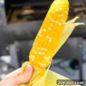 A hand grasps a grilled corn on the cob with husk in the background, subtly hinting at how to smoke corn on the cob for that rich, savory flavor.