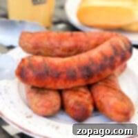 Smoked brats stacked on a white plate with tongs and buns in the background.