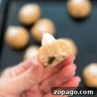 Close-up of a hand holding a half-eaten ball of edible raw cookie dough with a chocolate chip peeking out. Other dough balls and a baking tray are blurred in the background, tempting you with their gooey promise.
