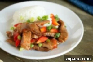 A plate of Chinese pepper steak, cooked to perfection in a pressure cooker, features tender beef stir-fry with red peppers, green onions, and a serving of fluffy white rice.