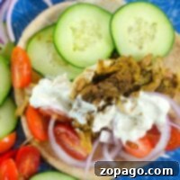 Plate with an instant pot chicken shawarma gyro sandwich, topped with sliced cucumbers, tomatoes, and onions, served alongside warm pita bread and a generous drizzle of sauce.
