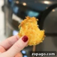Crunchy Air Fryer Zucchini Chips 5 Hand holding a bite of crispy breaded zucchini in front of an air fryer, nails painted red, showcasing the perfect snack alongside freshly made air fryer zucchini chips.