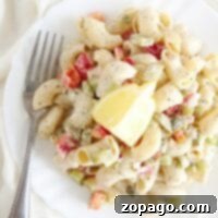 A white plate of dill pickle pasta salad with vegetables, topped with lemon wedges, and a fork on the side.
