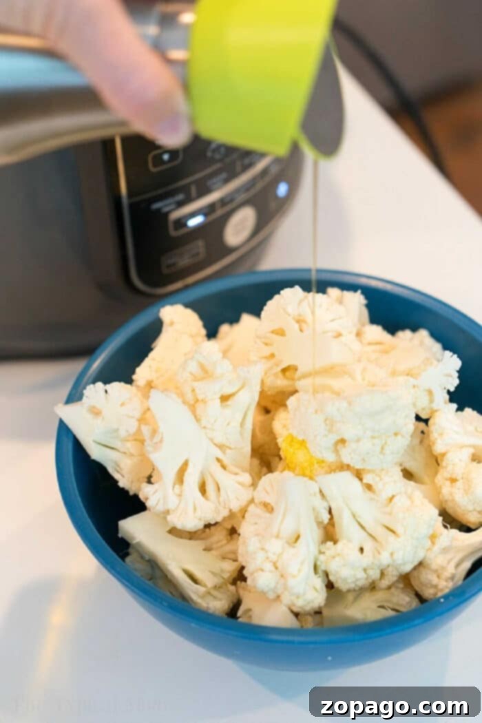Easy air fryer cauliflower is easy to make in your Ninja Foodi or other machine. Healthy and full of flavor it's a great vegetable side dish or snack. #airfryer #ninjafoodi #cauliflower #vegetables #vegetarian #sidedish Olive oil is being poured onto fresh cauliflower florets in a blue bowl, perfect for making crispy air fryer cauliflower near an electric cooker.
