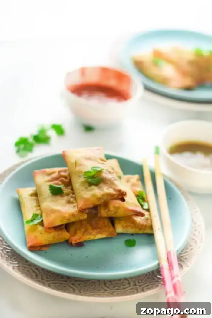 Discover the best air fryer egg rolls – a healthy and delicious appetizer or dinner. Perfect for your new kitchen appliance, whether you prefer chicken or vegetarian fillings. A plate with golden-brown air fryer egg rolls, garnished with fresh cilantro. Chopsticks rest beside the plate, and two bowls of dipping sauces, one reddish and one brown, are visible in the background on a clean white surface.