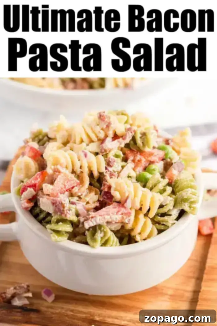 Smoky Bacon Pasta Perfection 2 A white bowl filled with a colorful bacon pasta salad, which includes rotini, bacon, peas, and diced red bell peppers, is set on a wooden surface. The words "Ultimate Bacon Pasta Salad" are displayed at the top in bold black letters.