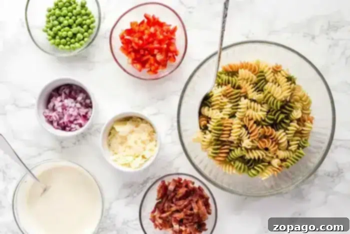 Smoky Bacon Pasta Salad 3 A variety of ingredients are laid out in small bowls on a white marble countertop. There is a large bowl of tri-color rotini pasta for the bacon pasta salad, peas, chopped red bell pepper, red onion, parmesan cheese, bacon pieces, and a bowl of creamy sauce. A spoon is in the pasta bowl.