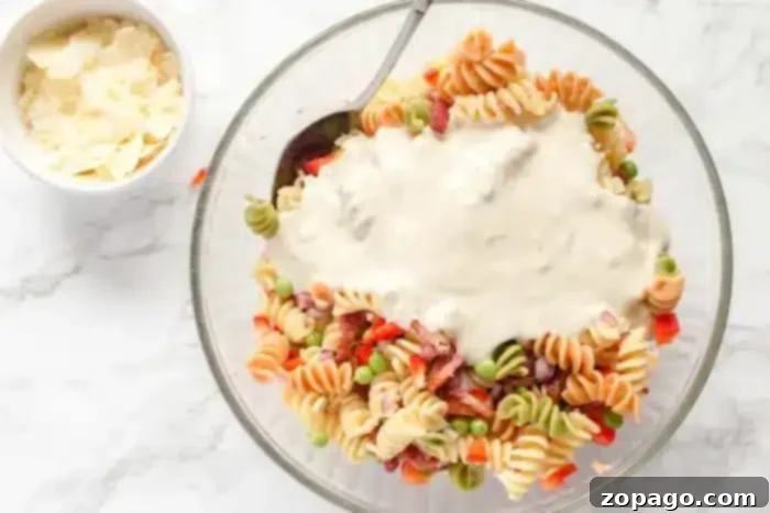 Smoky Bacon Pasta Salad 5 A clear glass bowl filled with colorful rotini pasta, peas, diced red bell peppers, and cubes of ham forms the heart of this enticing bacon pasta salad. A white creamy sauce is poured on top. A metal spoon is in the bowl. A small bowl beside it contains shaved Parmesan cheese.