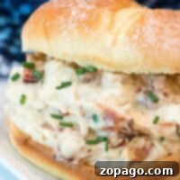 A sandwich filled with creamy Ninja Foodi crack chicken and bacon, garnished with chopped chives. It sits on a square, white plate, with a blurred blue patterned background.