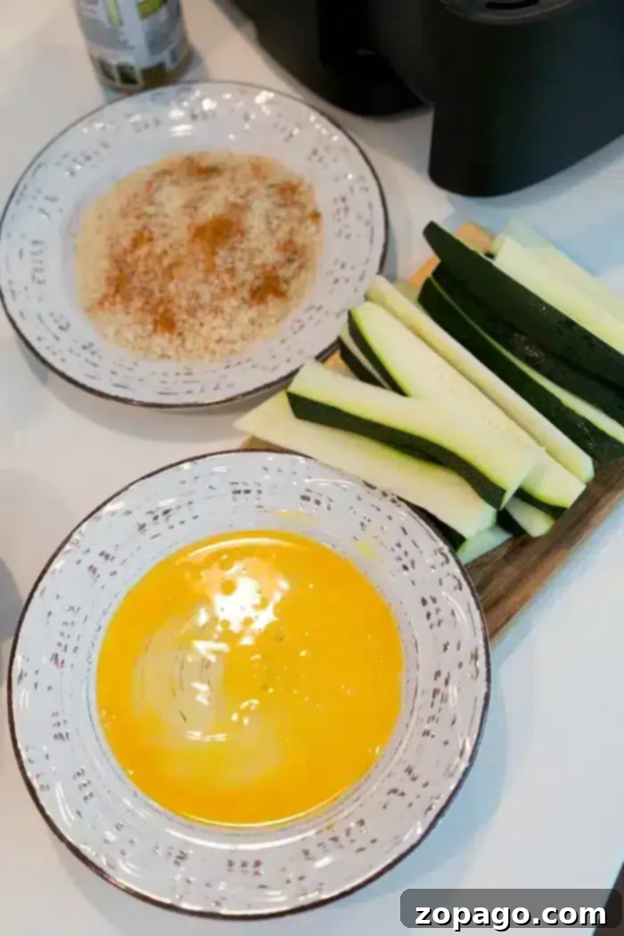 Best Air fryer zucchini fries are a healthy low carb snack your whole family will love! It's our favorite vegetable air fryer recipe. #airfryer #airfryerrecipes #zucchini #fries #zucchinifries #spears #lowcarb Two bowls on a white surface, one with whisked eggs, the other with a breadcrumb mixture. Next to the bowls are perfectly sliced zucchini on a cutting board, ready for breading to become delicious air fryer zucchini fries.