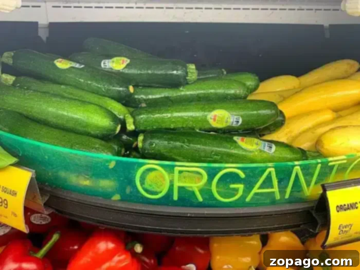 Crispy Air Fried Zucchini Sticks 6 A grocery store display features green zucchini with produce stickers and yellow squash, perfect for making air fryer zucchini fries. A partially visible yellow sign reads "ORGANIC." Red bell peppers are visible in the bottom of the frame.