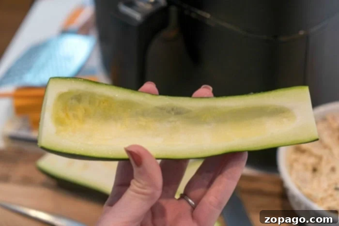 Stuffed Air-Fried Zucchini 3 A pair of hands meticulously scooping out the seeds and flesh from a halved zucchini using a spoon, demonstrating the preparation process for stuffed zucchini boats.