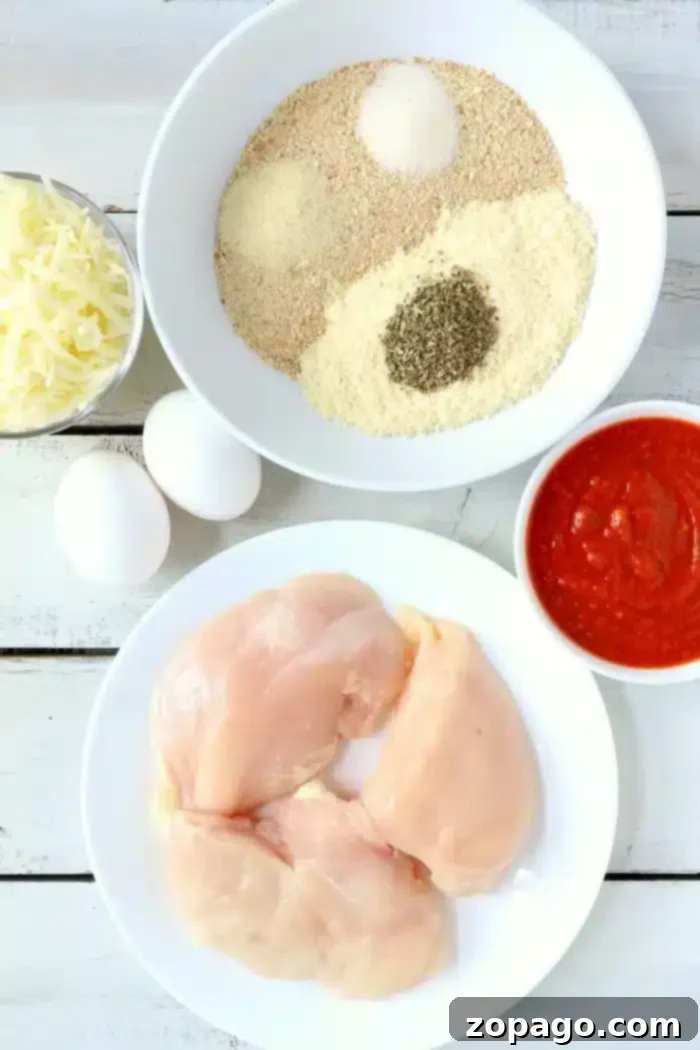 Ingredients for Air Fryer Chicken Parm: Chicken, Eggs, Breadcrumbs, Cheese, Marinara A flat lay of cooking ingredients for air fryer chicken parm: raw chicken breasts on a white plate, two eggs, shredded cheese, a bowl of breadcrumbs mixed with seasonings, and a bowl of red sauce, all placed on a white wooden surface.