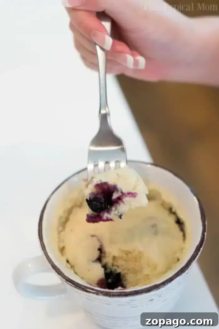 Blueberry Mug Bliss 7 A hand holding a fork with a piece of perfectly baked blueberry muffin in a mug, positioned above the ceramic mug on a white surface, highlighting its delightful texture and readiness to eat.