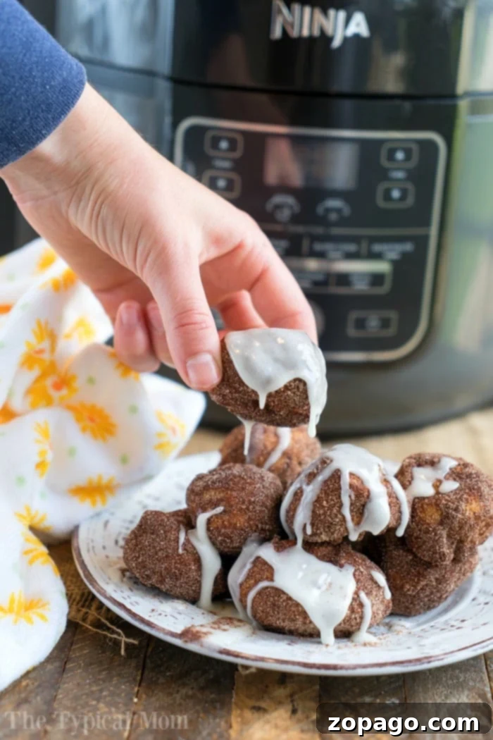 Easy air fryer donut recipe using cinnamon rolls and cinnamon and sugar! Soft on the inside and crispy outsides, make these in your Ninja Foodi tonight! #airfryer #airfryerrecipe #donuts #cinnamonrolls #ninjafoodi #ninjafoodirecipes #dessert A hand holds an iced air fryer donut made with cinnamon rolls, with the trusty ninja foodi in the background.