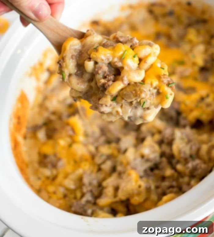 A hand holds a wooden spoon above a crockpot filled with a cheesy hamburger casserole. This savory delight combines macaroni, ground beef, and melted cheese, all garnished with fresh herbs for that perfect finish, making it a perfect family meal.