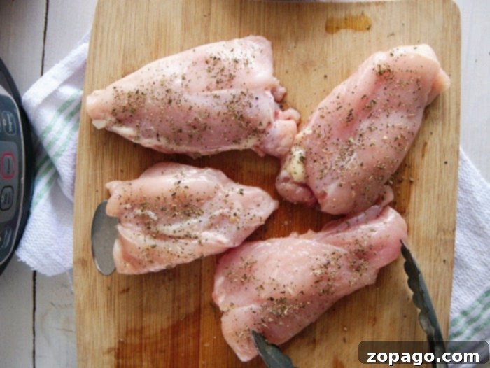 Slow Cooker Lemon Pepper Chicken 4 Four seasoned raw chicken thighs, ready for a crockpot lemon pepper chicken recipe, resting on a wooden cutting board next to a trusty pair of tongs.