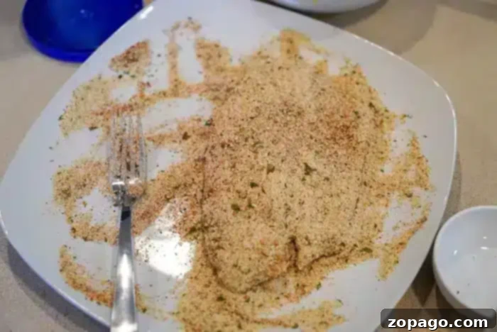 Crispy Air Fryer Tilapia 5 A piece of breaded tilapia fish is covered in a seasoned breadcrumb mixture on a white plate. A silver fork rests on the side of the plate, and there is a small empty white bowl and a blue lid in the background.