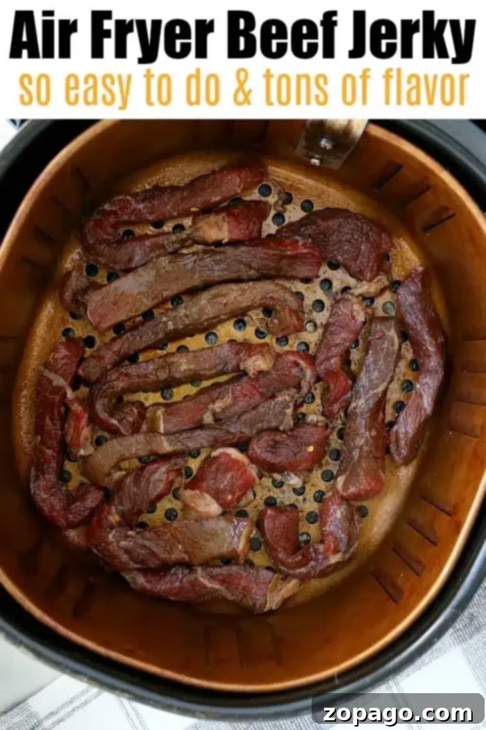 Best air fryer beef jerky is here! Ready to make tender beef in your Ninja Foodi or electric air fryer? We have an easy way to try your first batch. #airfryerbeefjerky #airfryer #airfryerrecipes #ninjafoodirecipes #foodi #ninjafoodi #thetypicalmom A metal air fryer basket is filled with pieces of savory beef jerky. The top of the image contains text that reads: "Air Fryer Beef Jerky so easy to do & tons of flavor." The setup is on a gray-patterned countertop.