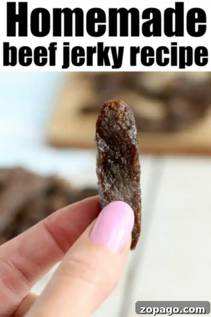 Homemade beef jerky is here! Ready to make tender beef in your Ninja Foodi or electric air fryer? We have an easy way to try your first batch. #airfryerbeefjerky #airfryer #airfryerrecipes #ninjafoodirecipes #foodi #ninjafoodi #thetypicalmom A close-up of a hand with pink-painted nails holding a piece of beef jerky. "Homemade air fryer beef jerky recipe" is written in bold black letters at the top of the image. In the background, more pieces of jerky are spread out on a wooden surface.