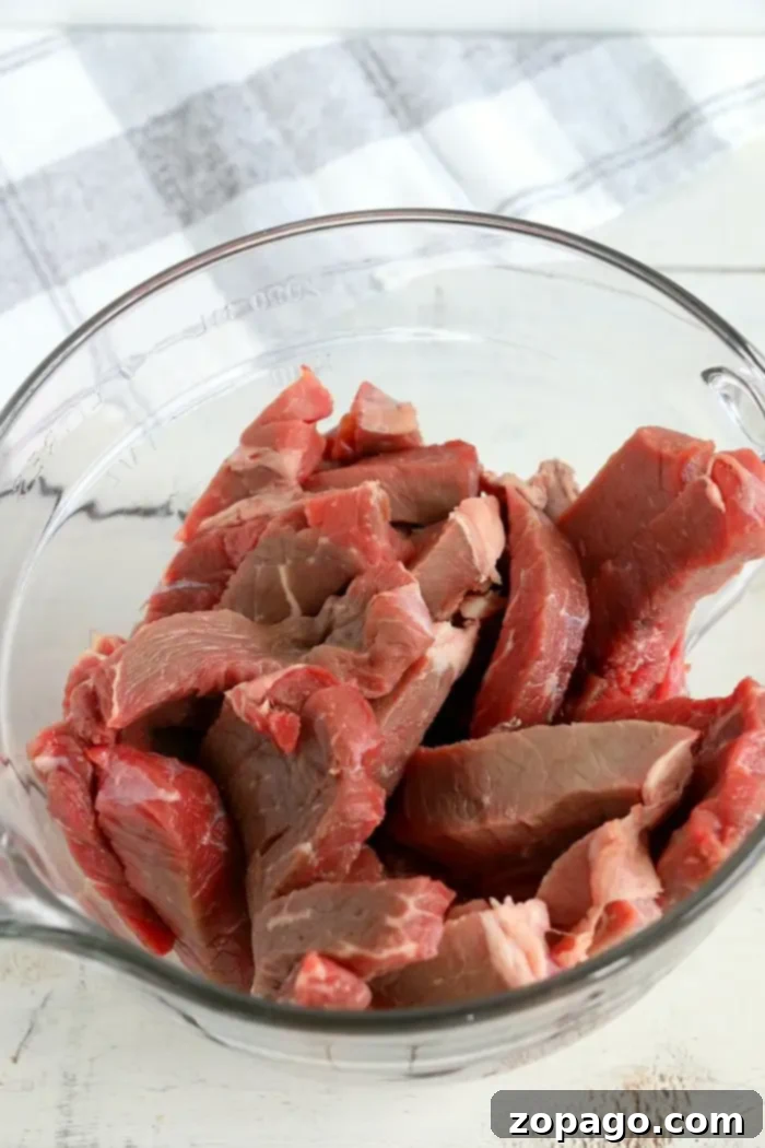 How to make beef jerky is here! Ready to make tender beef in your Ninja Foodi or electric air fryer? We have an easy way to try your first batch. #airfryerbeefjerky #airfryer #airfryerrecipes #ninjafoodirecipes #foodi #ninjafoodi #thetypicalmom A clear glass bowl filled with raw, thinly sliced beef pieces, perfect for making air fryer beef jerky.